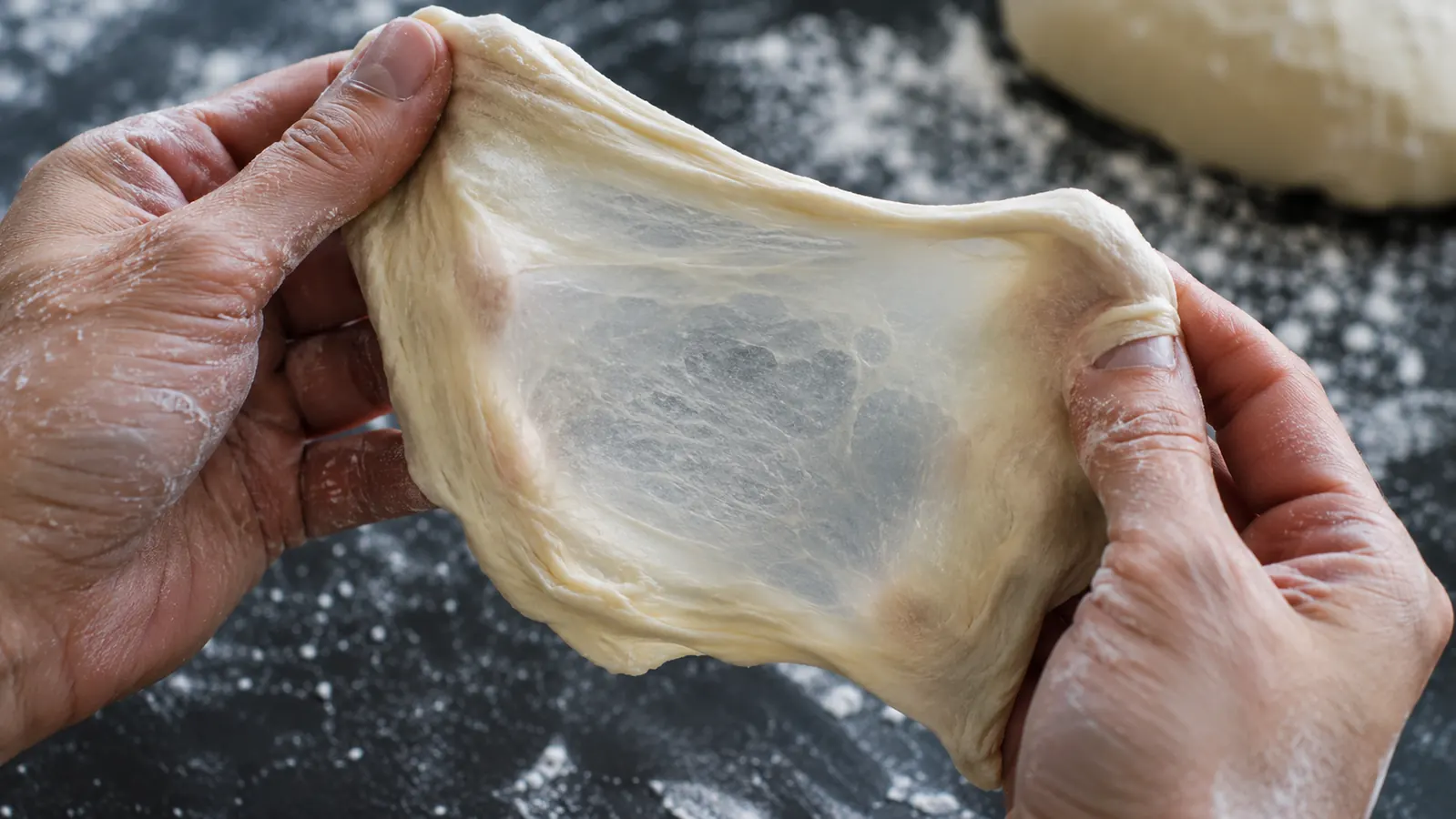 Hands stretching a small piece of dough into a thin translucent sheet on a floured surface
